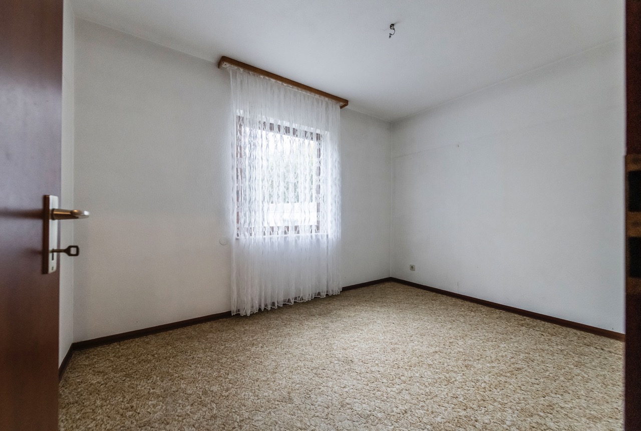 Leeres Zimmer mit Teppichboden, weißer Wand und Fenster mit Gardine.