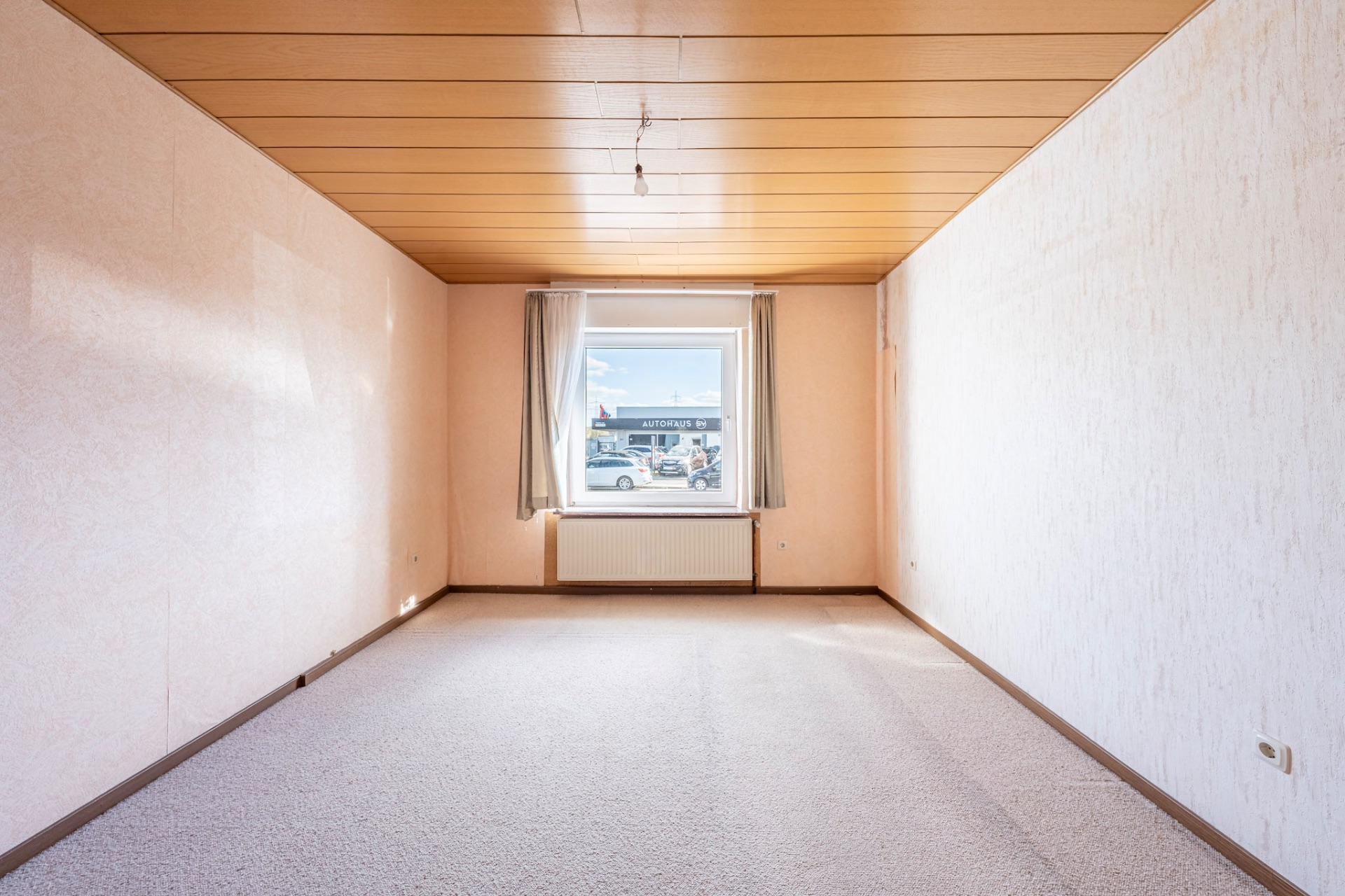 Leeres Zimmer mit Teppichboden, Holzdecke und Fenster mit Vorhängen in der Mitte.
