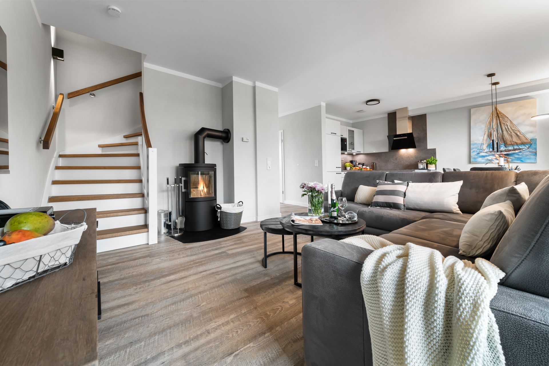 Modernes Wohnzimmer mit Sofa, Kaminofen, Esstisch und offener Küche, Treppe im Hintergrund.