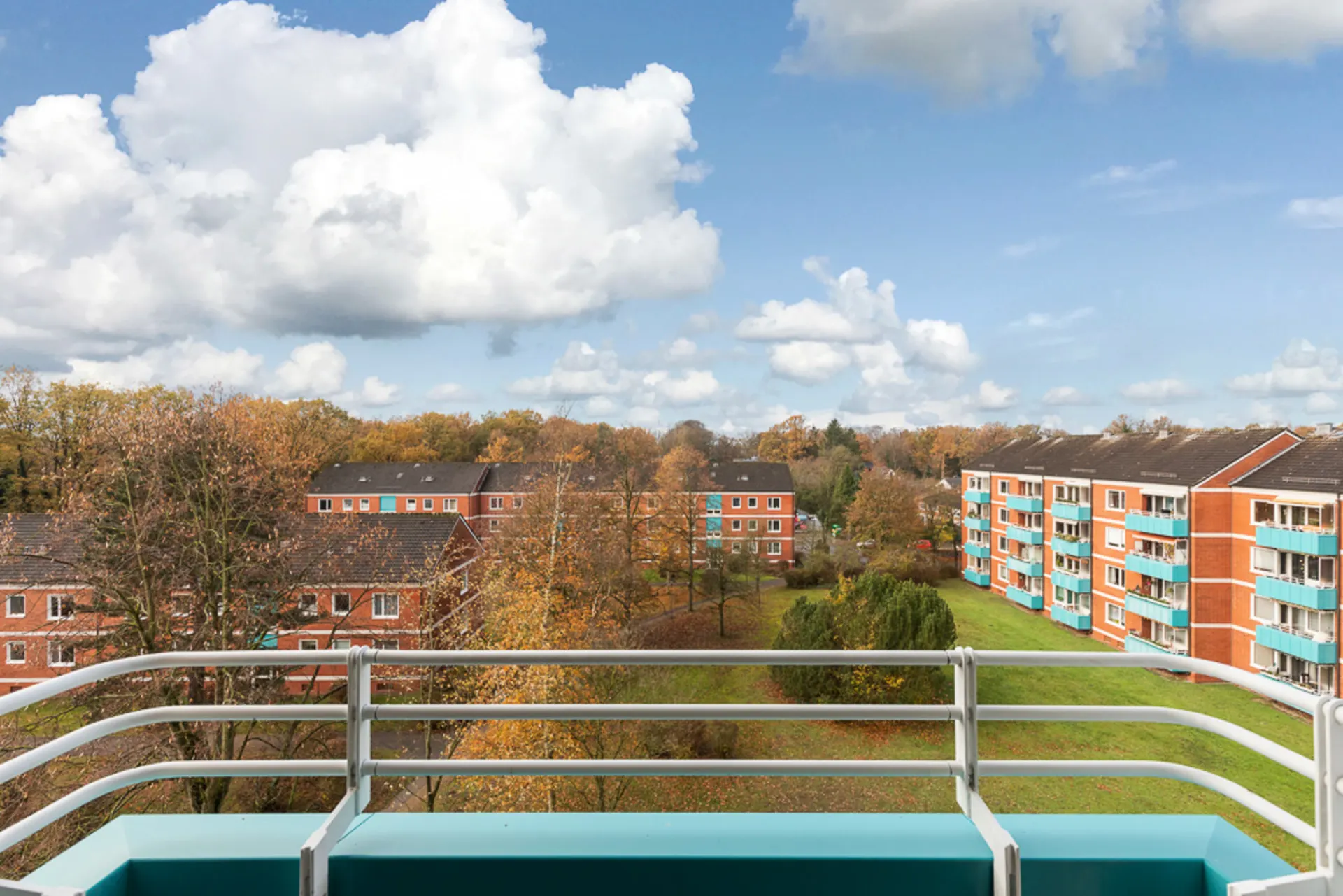 Blick vom Balkon auf grüne Wiese und rote Wohnhäuser mit Balkonen bei bewölktem Himmel.