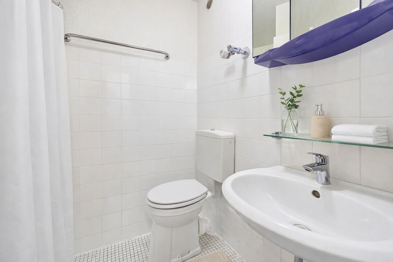 Helles Bad mit Toilette, Waschbecken, Glasablage und Handtüchern, weiße Fliesen.