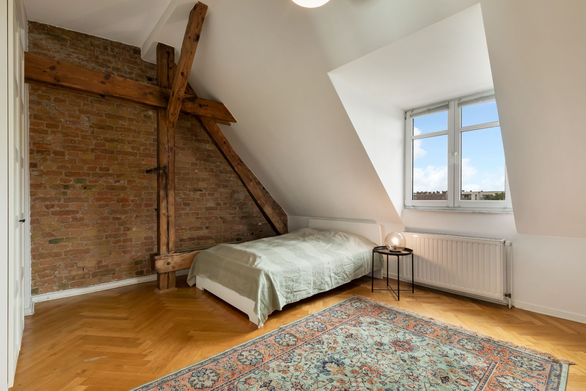 Schlafzimmer mit Holzbalken, Backsteinwand, Bett, Fenster, rundem Tisch und Teppichboden.