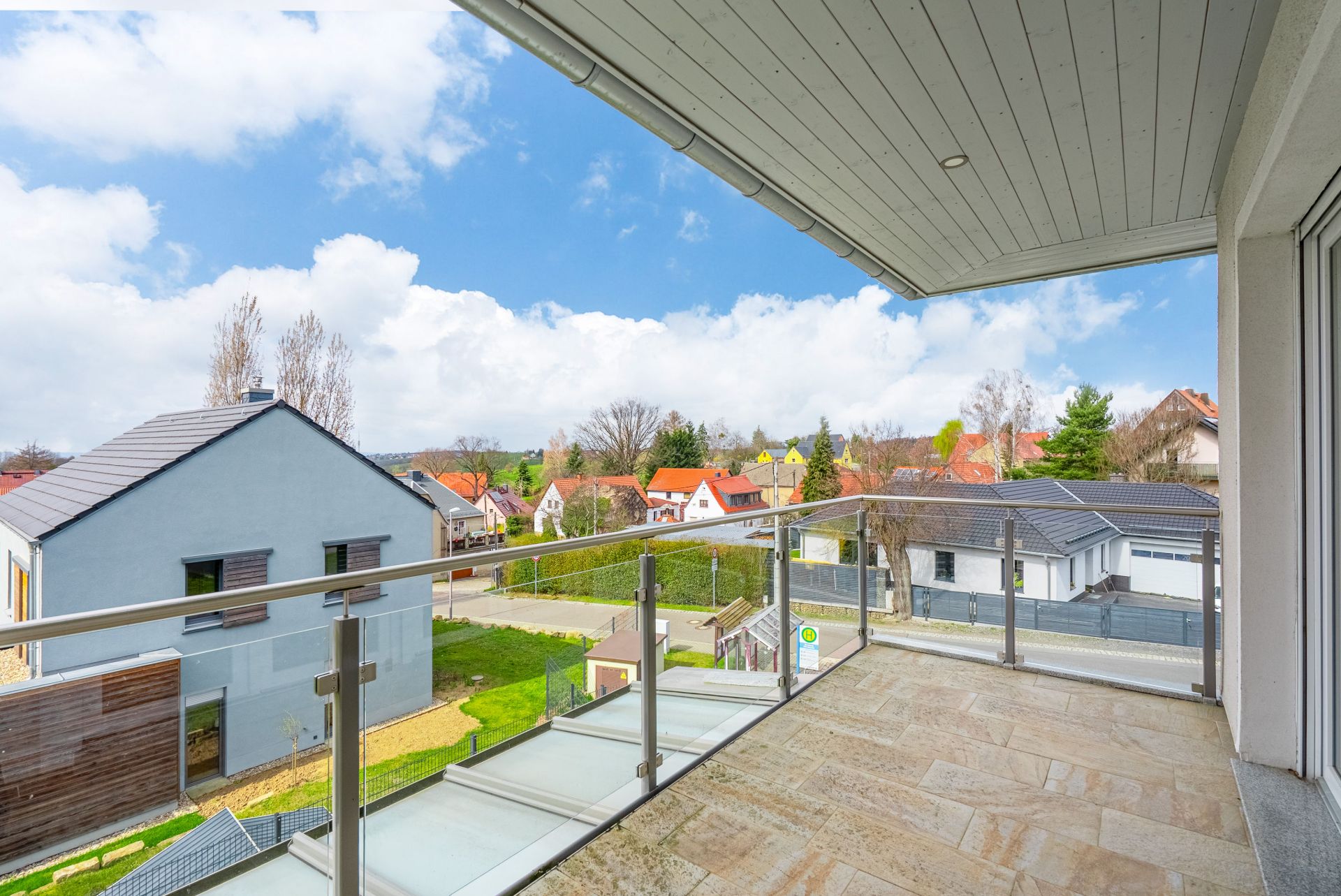 Balkon mit Glasgeländer und Blick auf Häuser und Gärten bei klarem Himmel.