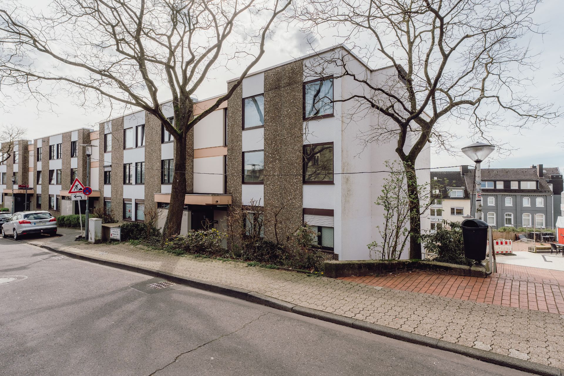 Mehrfamilienhaus mit drei Etagen und großen Fenstern an einer Straße mit Gehweg und Bäumen.