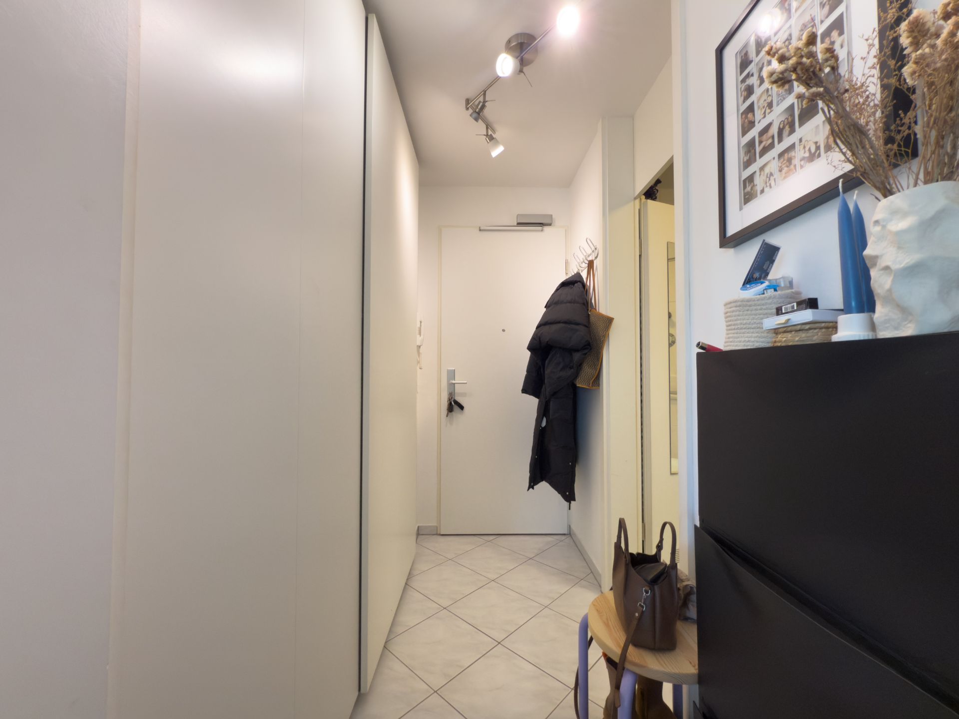 Schmaler Flur mit Garderobe, weißen Wänden, Kommode, Haken mit Jacken und heller Eingangstür.