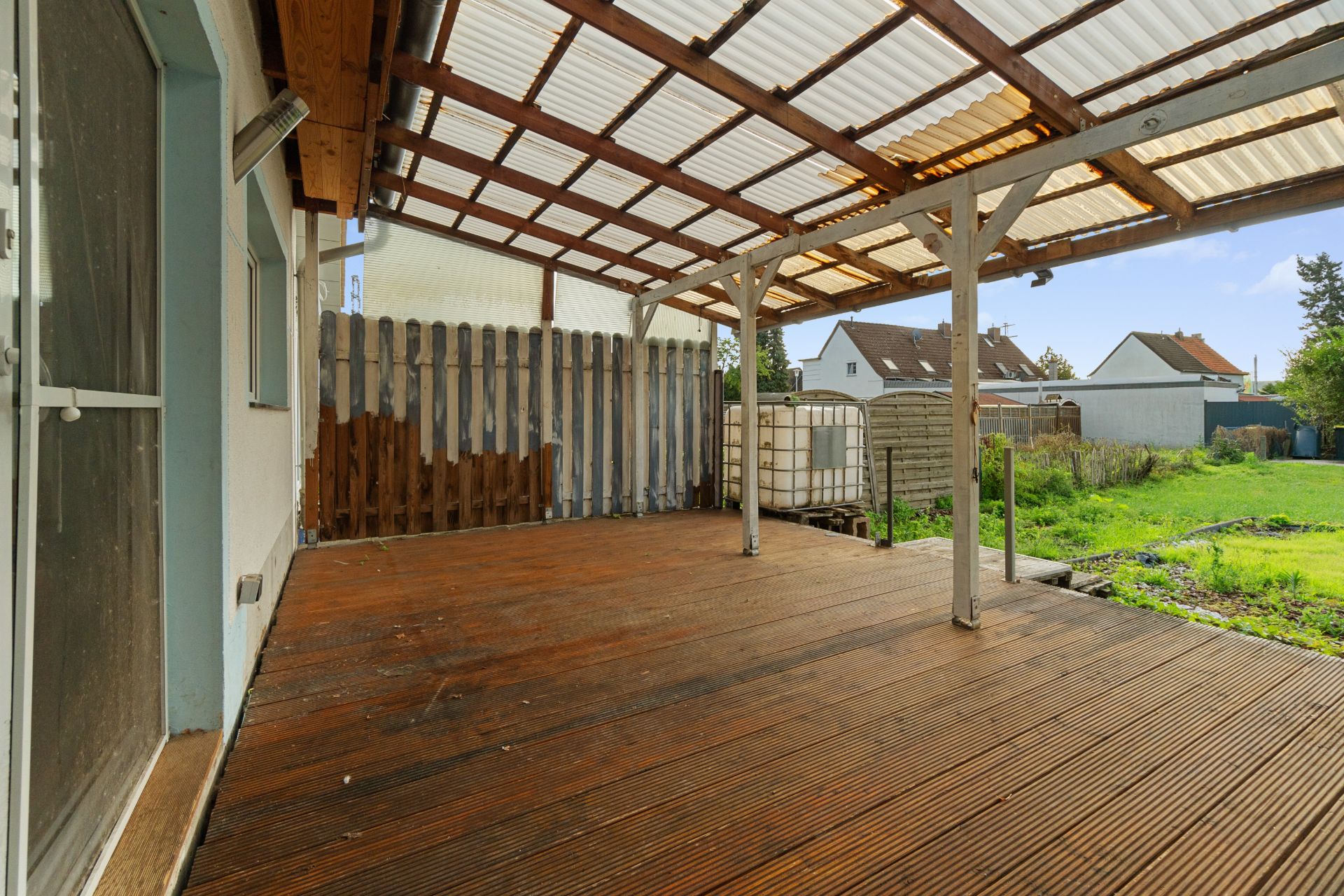 Überdachte Holzterrasse mit Blick auf einen Garten und angrenzenden Zaun.