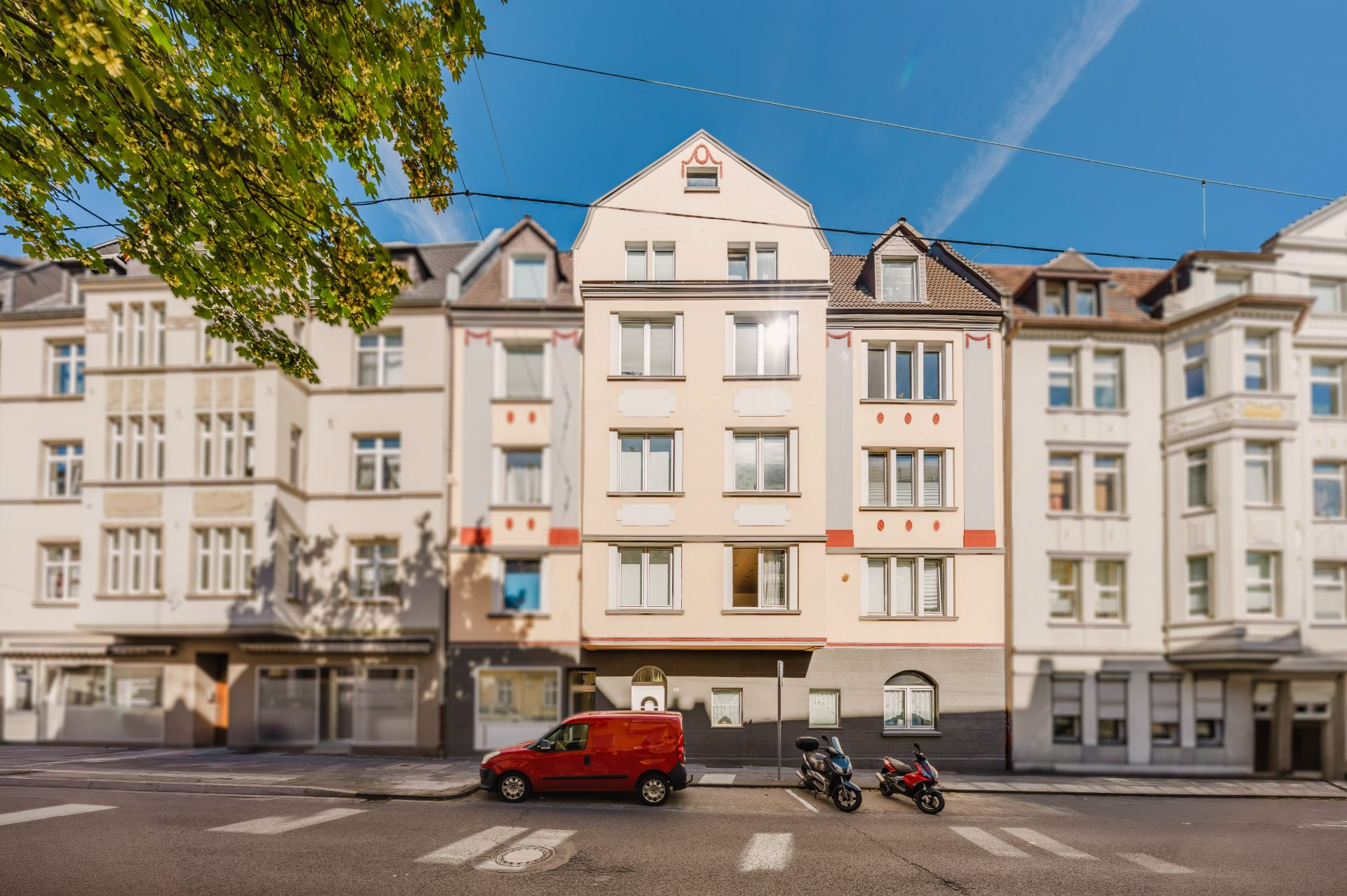 Mehrstöckiges Wohnhaus mit geparktem rotem Auto und zwei Motorrollern davor.