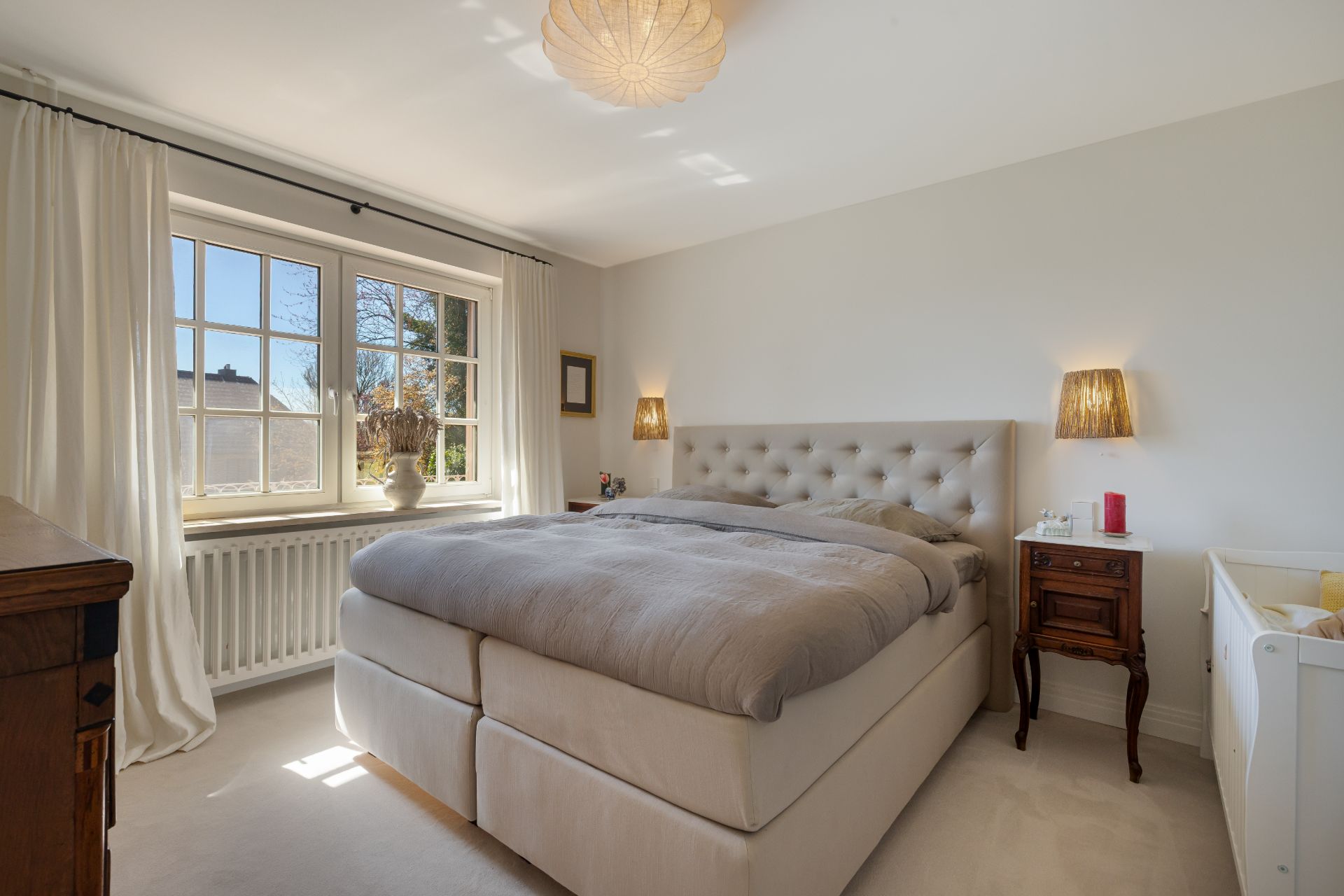 Helles Schlafzimmer mit großem Bett, Nachttischen, Fenster mit Vorhängen und Deckenleuchte.