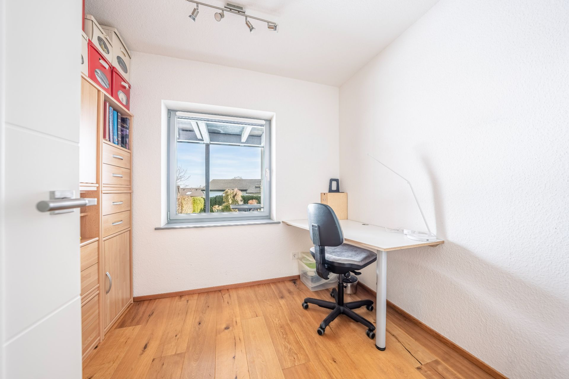 Helles Arbeitszimmer mit Schreibtisch, Bürostuhl, Fenster und Holzregal an der Wand.