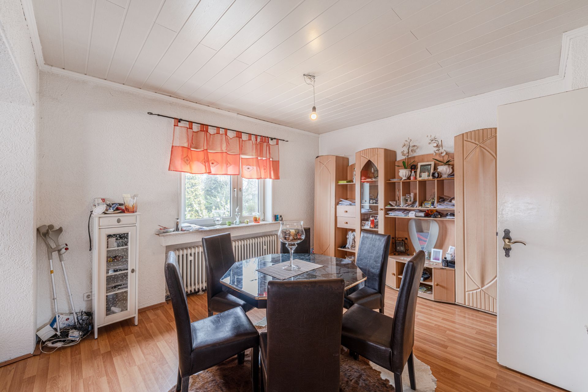 Wohnzimmer mit Esstisch, fünf Stühlen, Regalwand und Fenster mit orangefarbener Gardine.