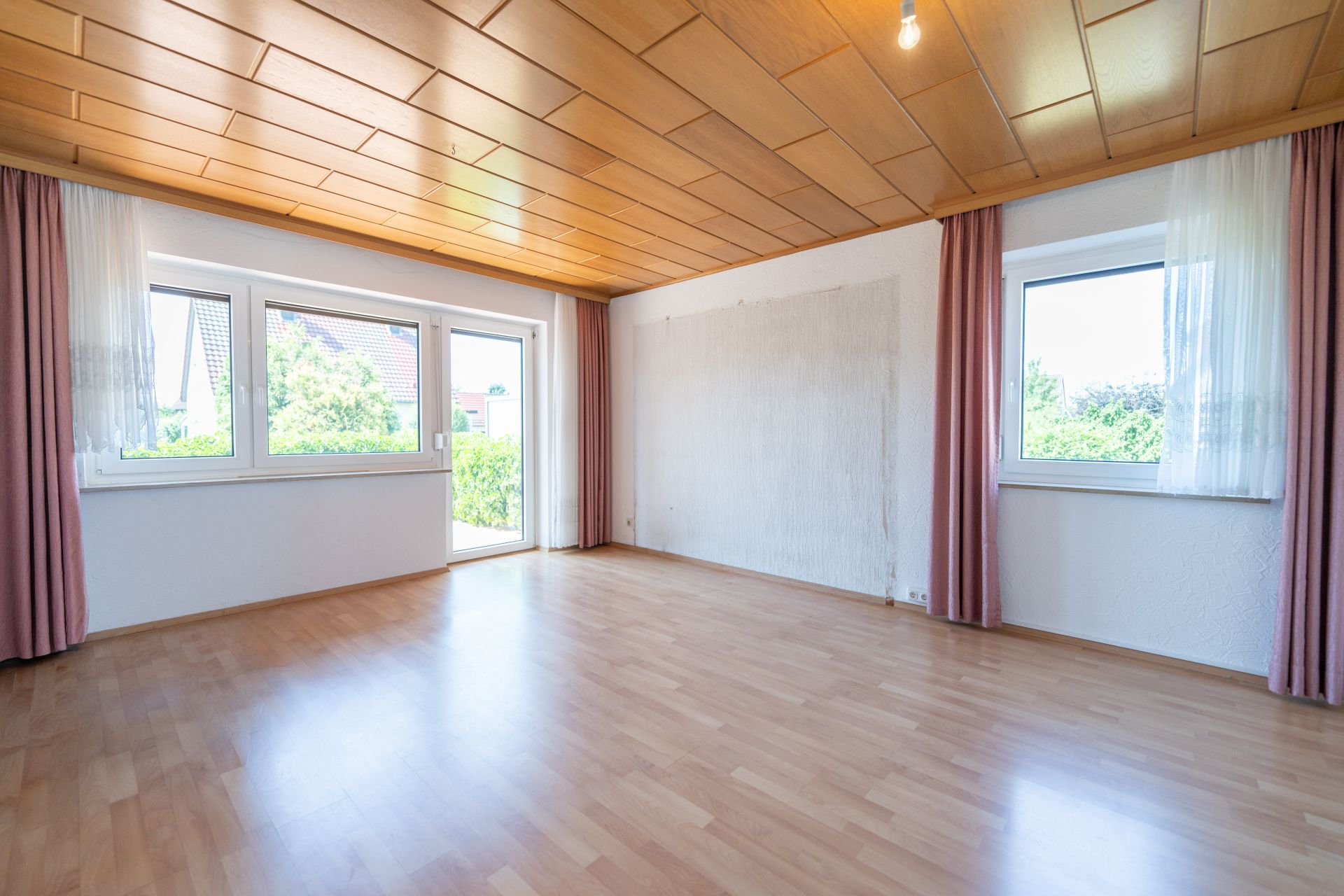Helles, leeres Zimmer mit Holzdecke, großen Fenstern und Vorhängen, Parkettboden.