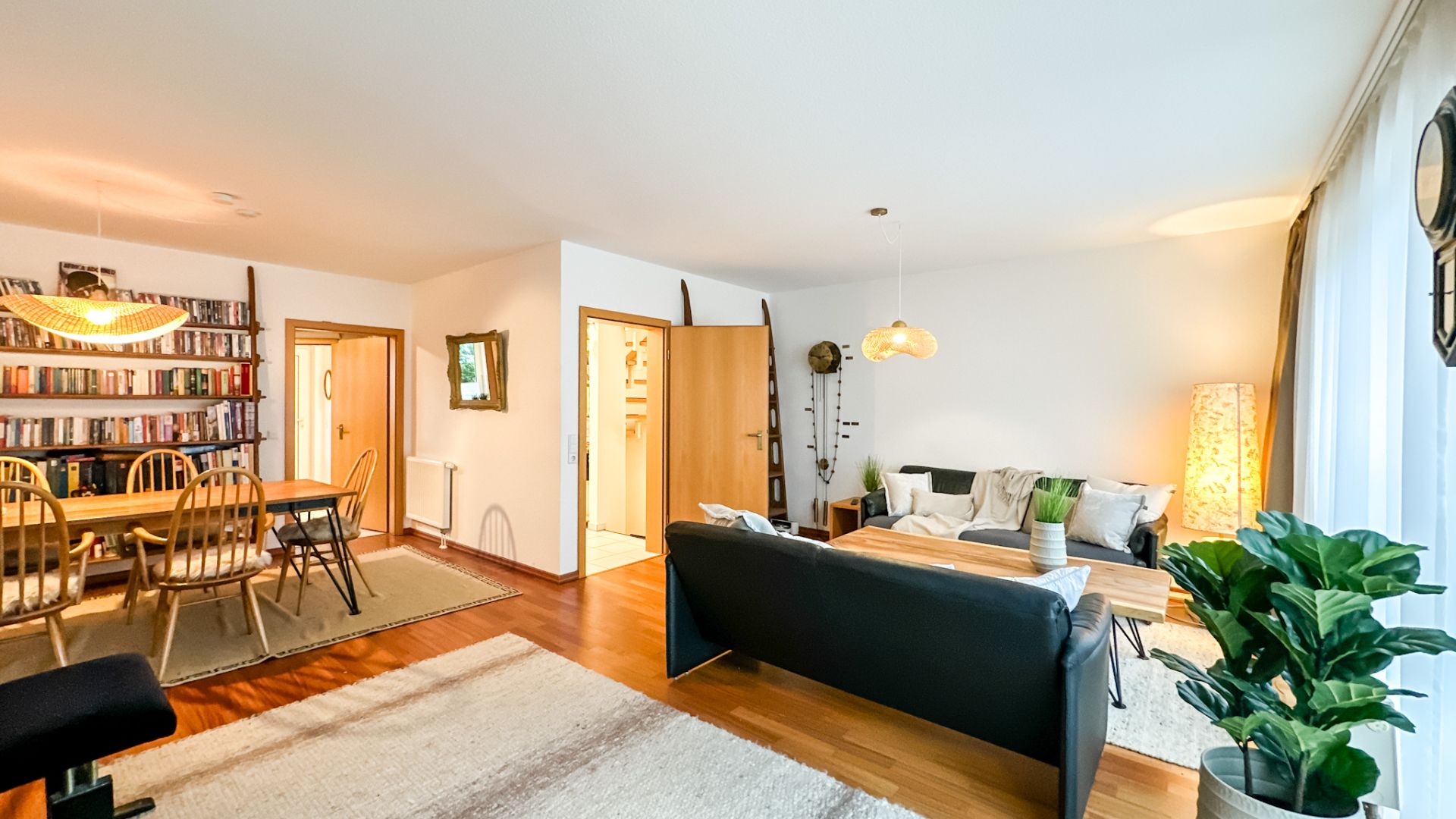 Helles Wohnzimmer mit Sofa, Esstisch, Bücherregal und großen Fenstern mit Vorhängen.