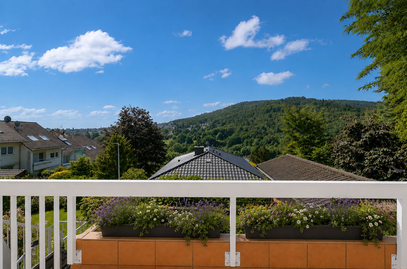 Blick von einem Balkon auf Gärten, Nachbarhäuser und bewaldete Hügel im Hintergrund.