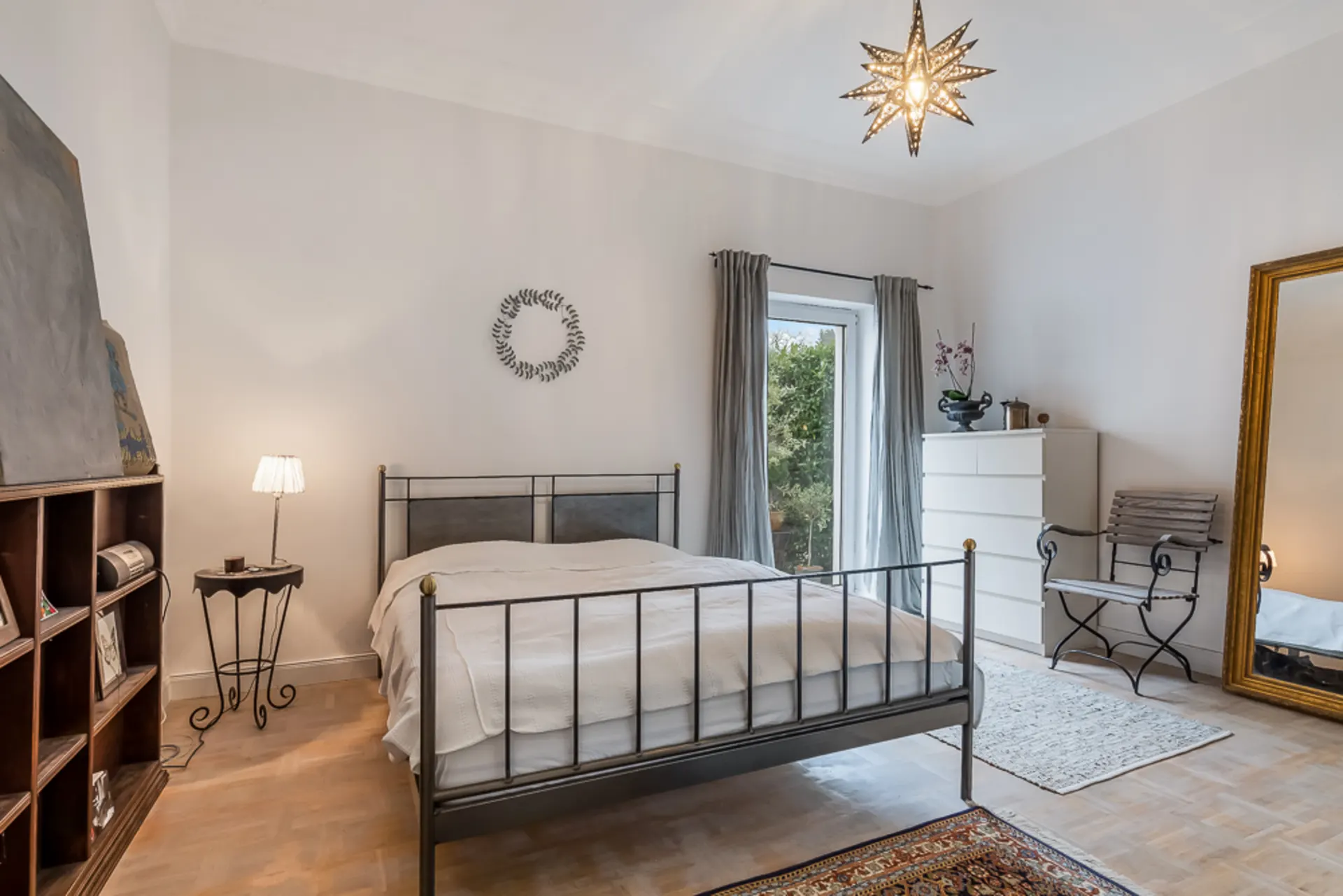 Helles Schlafzimmer mit Metallbett, großem Spiegel, Kommode und Fenster mit Blick ins Grüne.