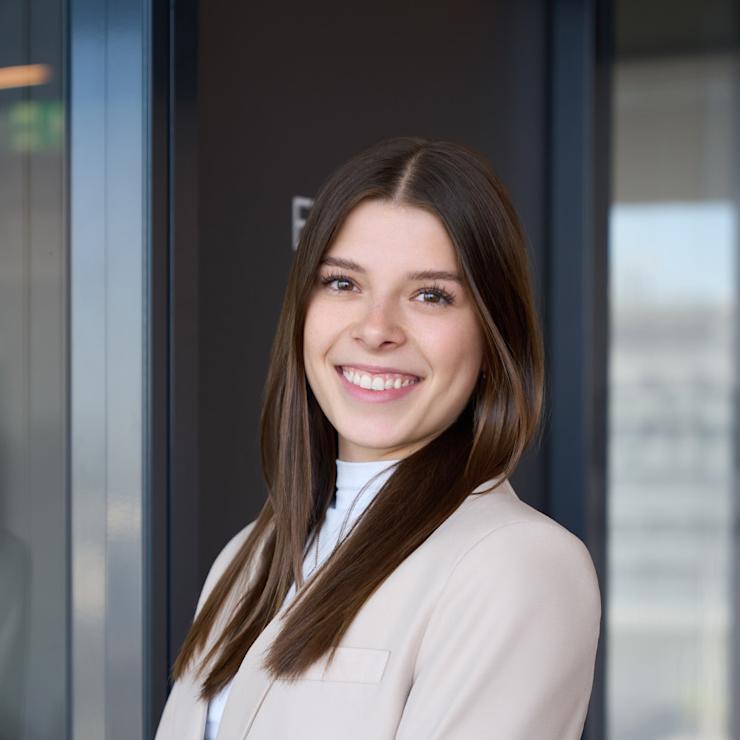 Junge Frau mit langen braunen Haaren und beigem Blazer steht vor einer Glastür.