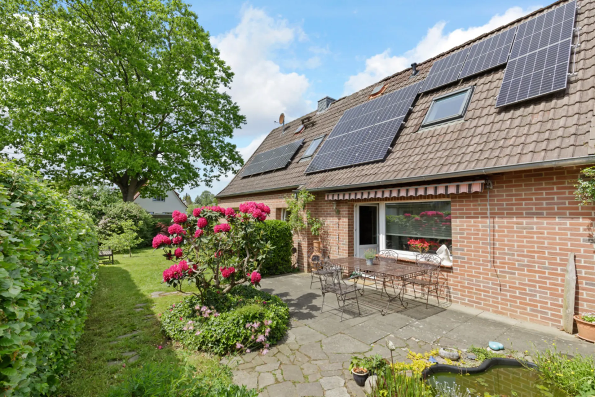 Backsteinhaus mit Solarpanelen, Terrasse, Gartentisch, Blumen und kleinem Teich im Garten.