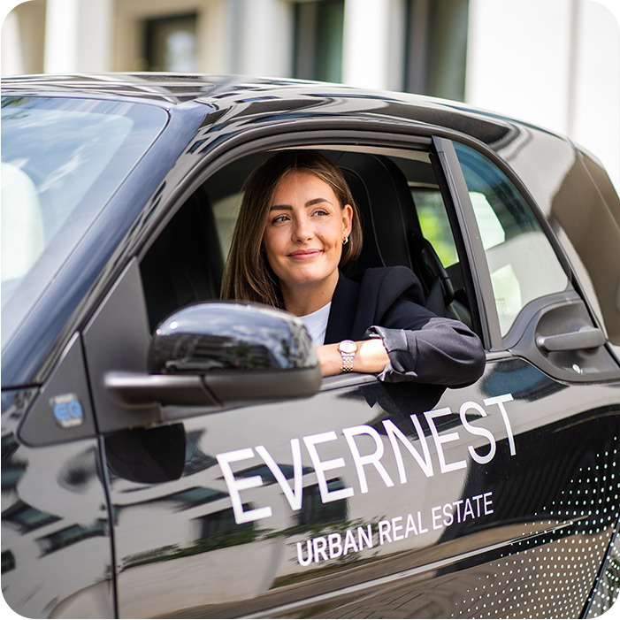 Frau sitzt im Auto mit der Aufschrift 'EVERNEST URBAN REAL ESTATE' vor Wohngebäuden.