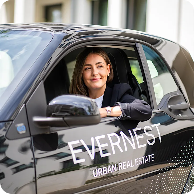 Frau sitzt im Auto mit der Aufschrift 'EVERNEST URBAN REAL ESTATE' vor Wohngebäuden.