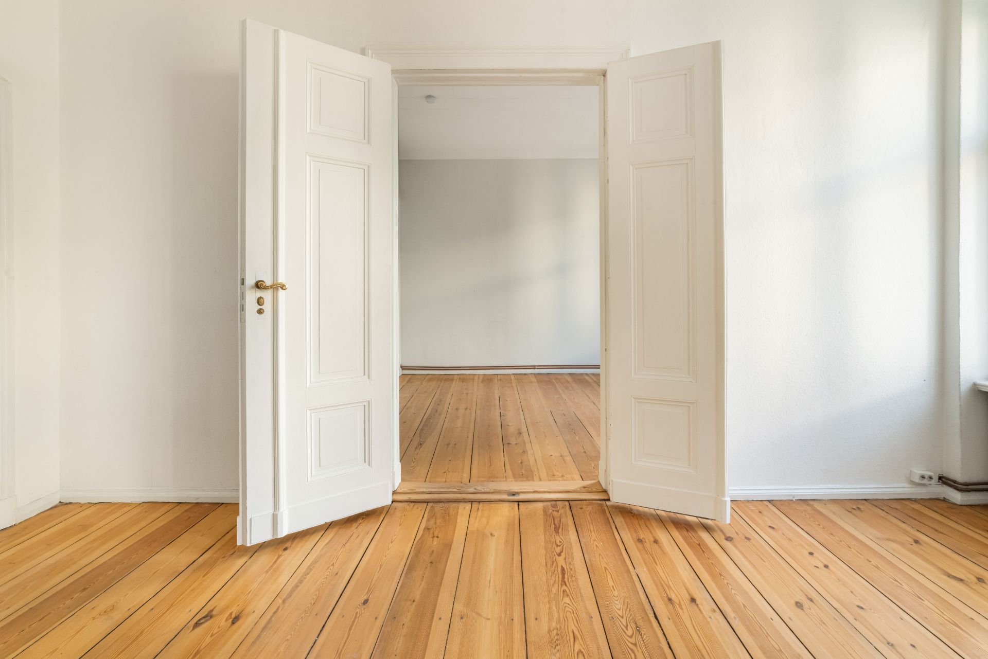Helles Zimmer mit Holzboden und offenen weißen Doppeltüren, leere Wände.