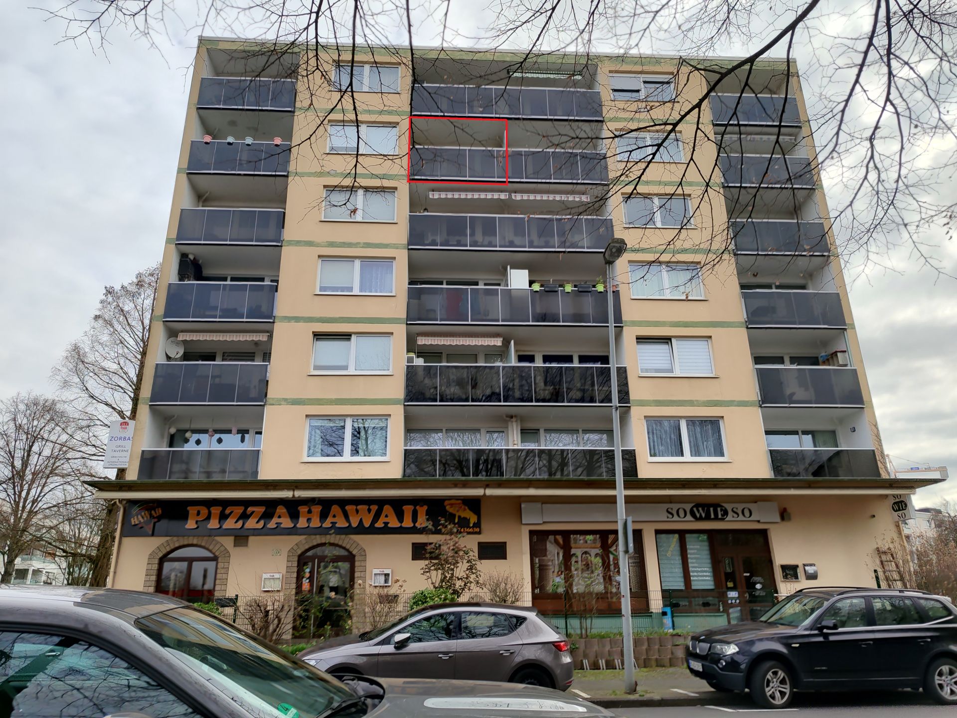 Mehrstöckiges Wohnhaus mit Balkonen, im Erdgeschoss befinden sich zwei Geschäfte, davor parken Autos.