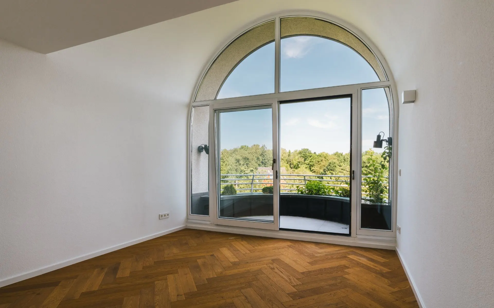 Leerer Raum mit großem Rundbogenfenster und Blick auf einen Balkon und Bäume.