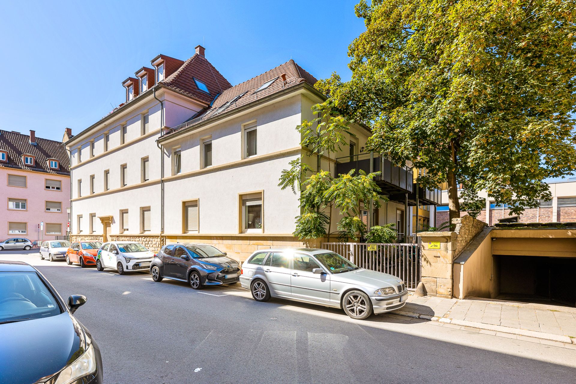 Wohnhaus mit mehreren Stockwerken, geparkte Autos davor, Bäume und Garageneinfahrt rechts.