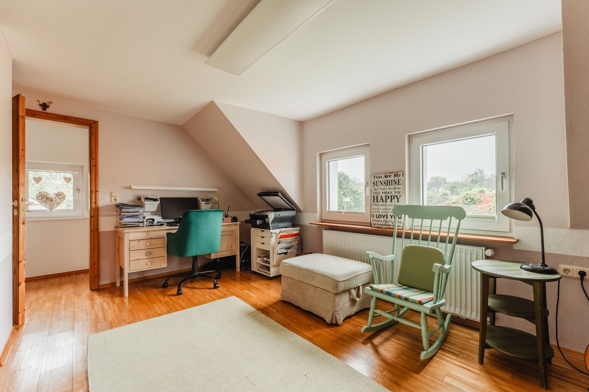Helles Arbeitszimmer mit Schreibtisch, grünem Stuhl, Sessel, Schaukelstuhl und zwei Fenstern.
