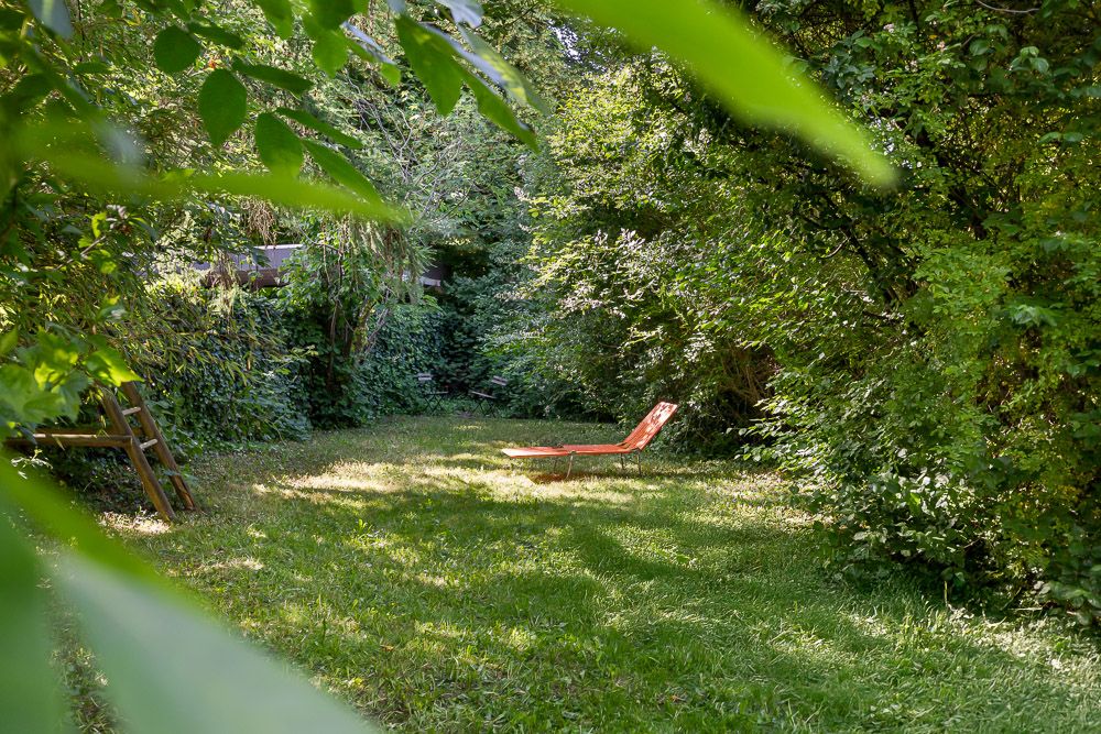 Grüner Garten mit einer roten Sonnenliege in der Mitte, umgeben von Bäumen und Büschen.