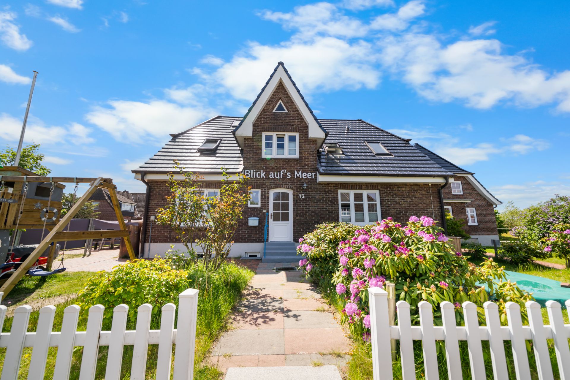 Backsteinhaus mit Garten, Blumen und Spielplatz, blauer Himmel im Hintergrund.