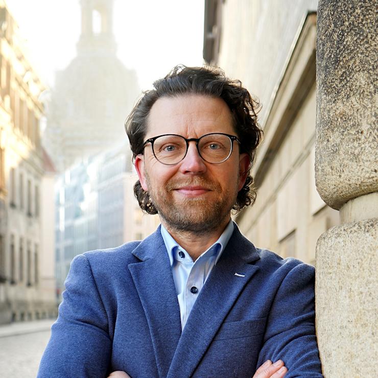 Mann mit Brille und blauem Sakko steht auf einer gepflasterten Straße vor einem Gebäude.