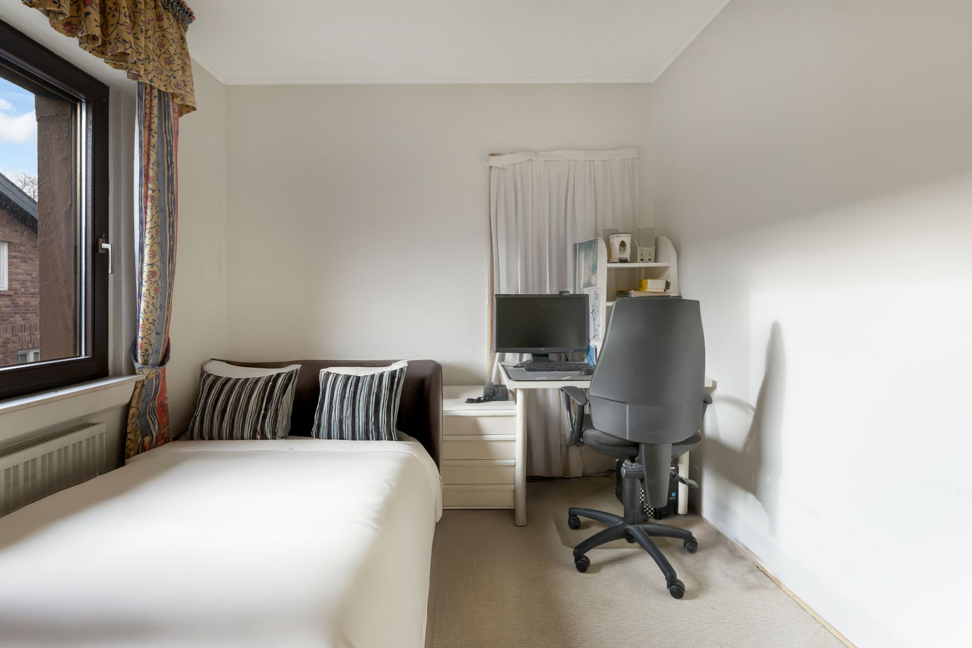 Kleines Zimmer mit Bett, Kissen, Schreibtisch, Monitor und Bürostuhl am Fenster.