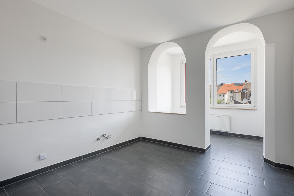 Leerer, heller Raum mit grauem Fliesenboden, weißen Wänden und zwei großen Fenstern.