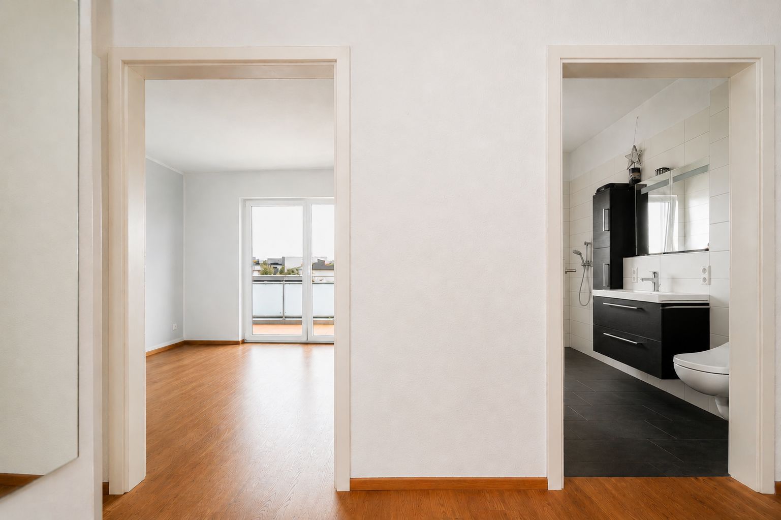 Blick vom Flur in ein Wohnzimmer mit Balkon und in ein modernes Badezimmer mit Waschbecken und Toilette.
