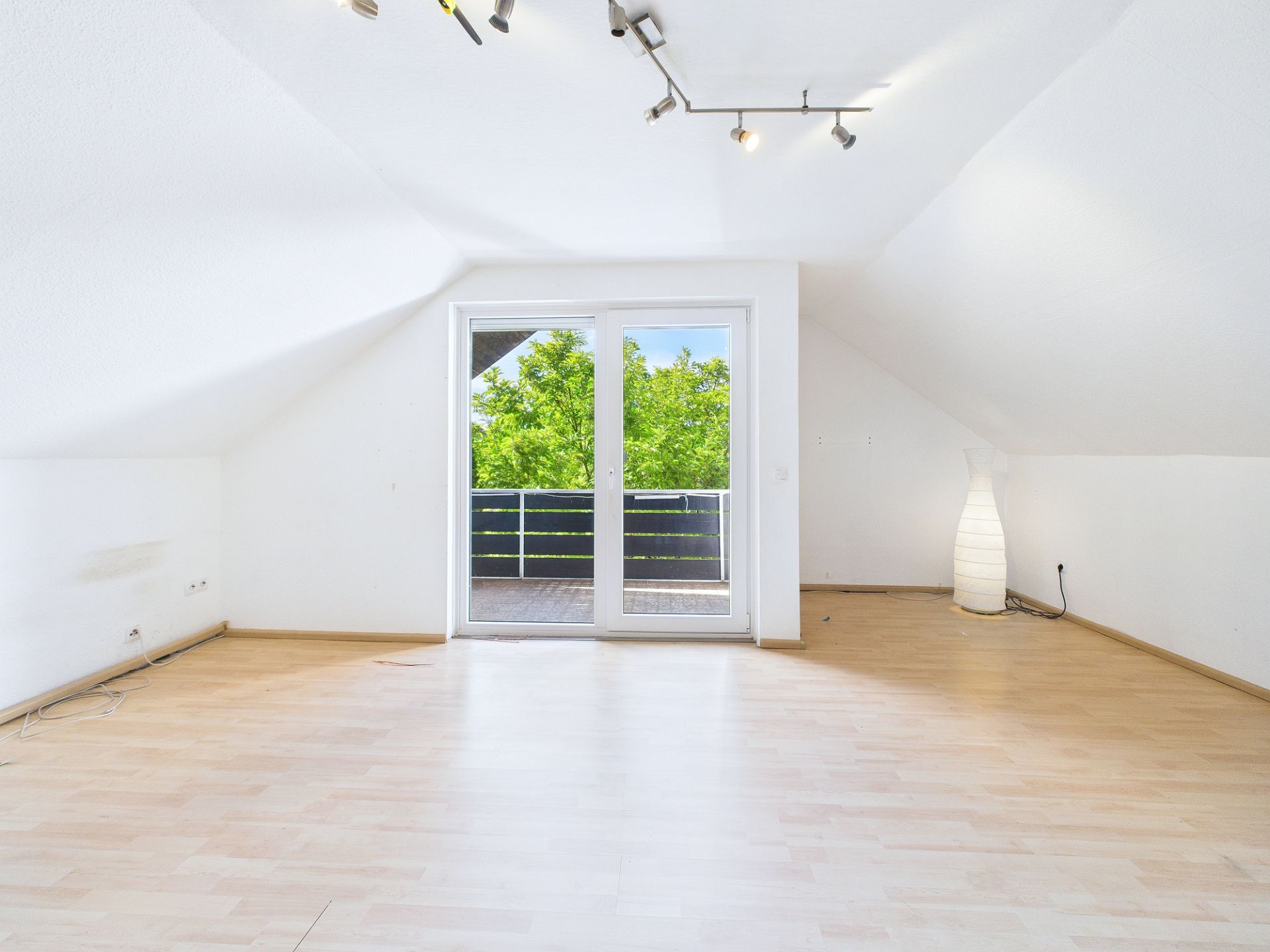 Helles Zimmer mit Dachschräge, Parkettboden, Glastür zum Balkon und Stehlampe in der Ecke.