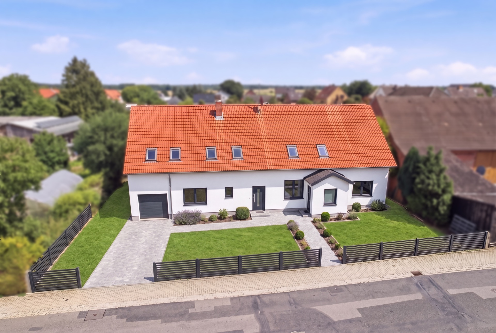 Modernes Einfamilienhaus mit rotem Dach, gepflegtem Garten und Einfahrt, umzäunt an einer Straße.