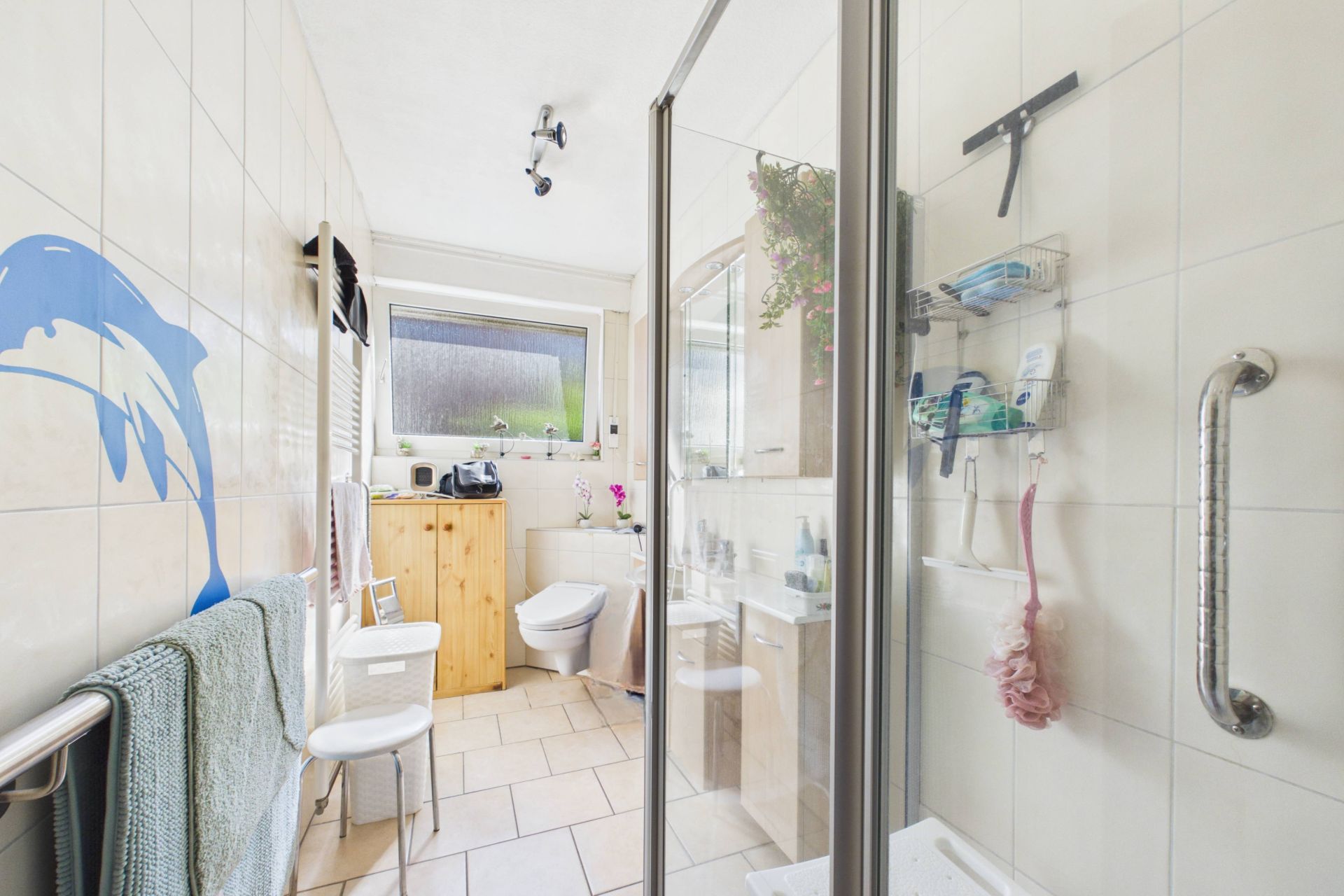 Helles Badezimmer mit Dusche, WC, Waschbecken, Holzschrank und Delfin-Wandbild.