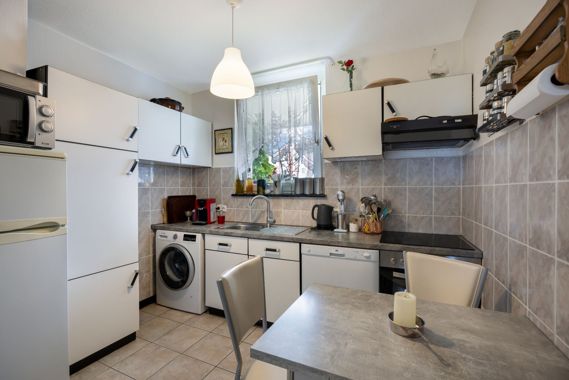 Kleine Küche mit weißen Schränken, Esstisch, Waschmaschine und Fenster mit Gardine.