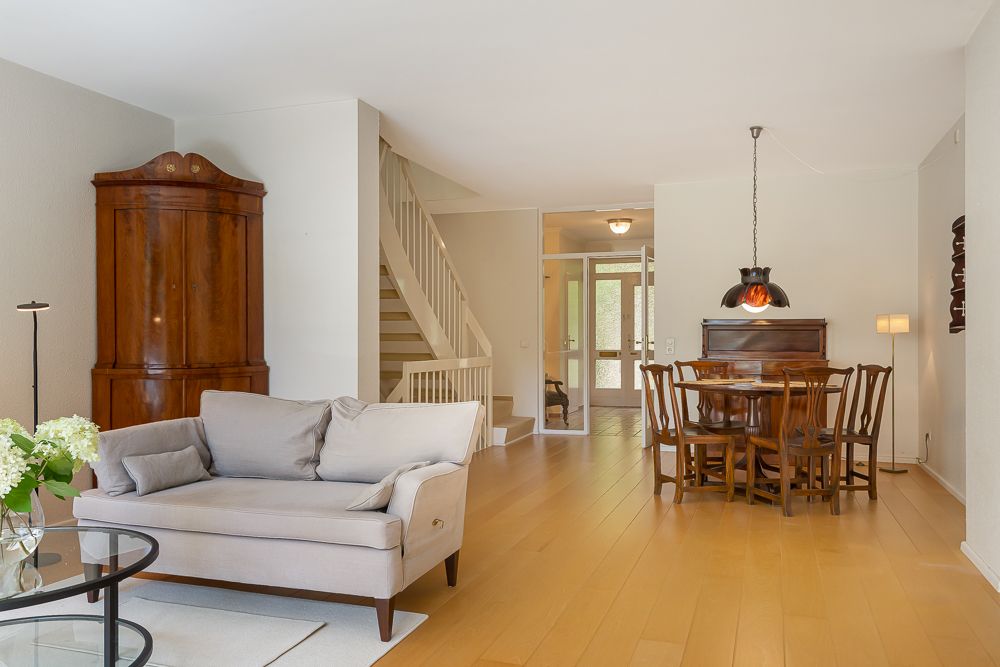Helles Wohnzimmer mit Sofa, Holztisch, Stühlen, Vitrine und einer Treppe im Hintergrund.