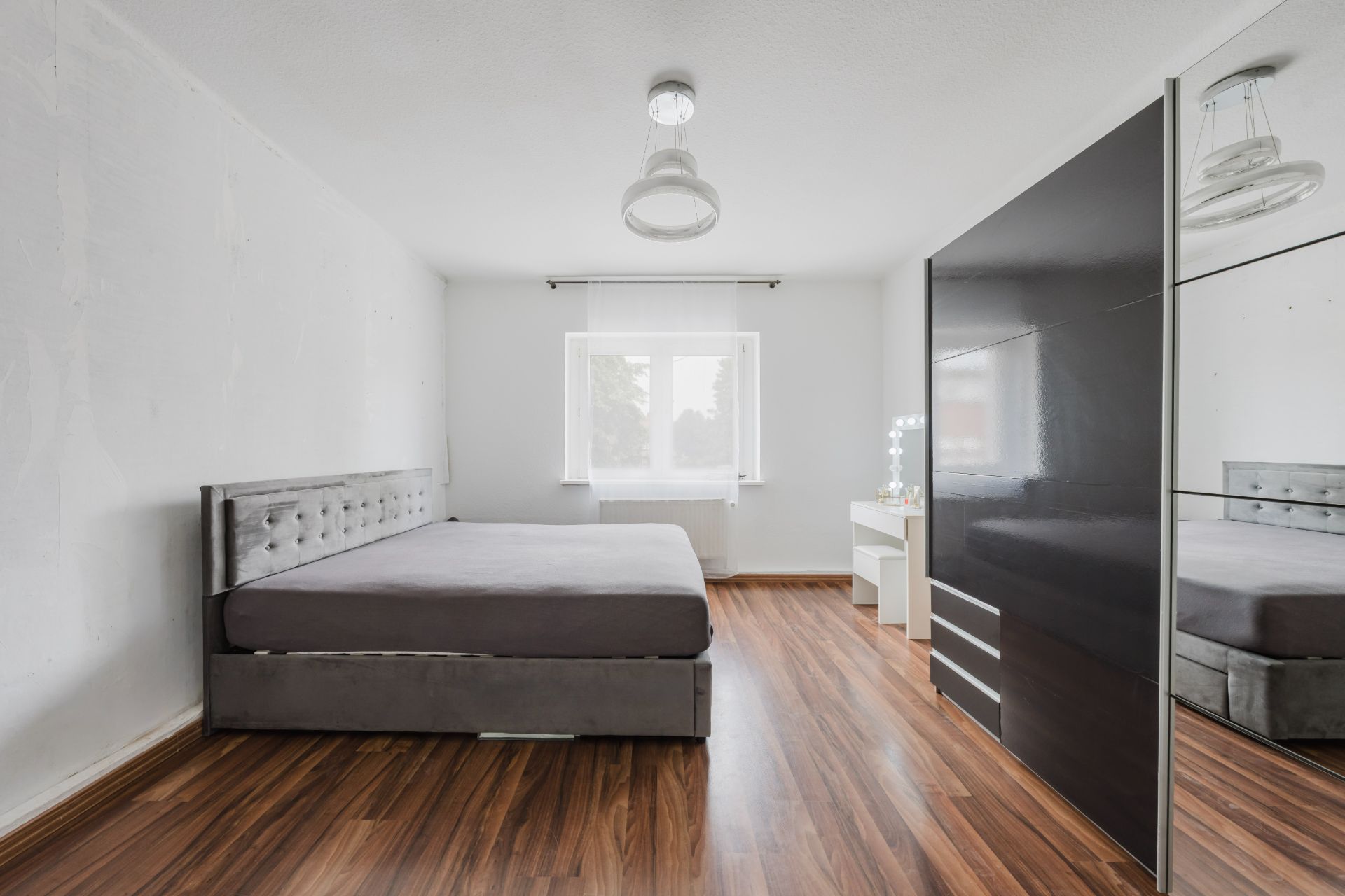 Schlafzimmer mit grauem Bett, großem Kleiderschrank, Holzfußboden und Fenster mit Vorhang.