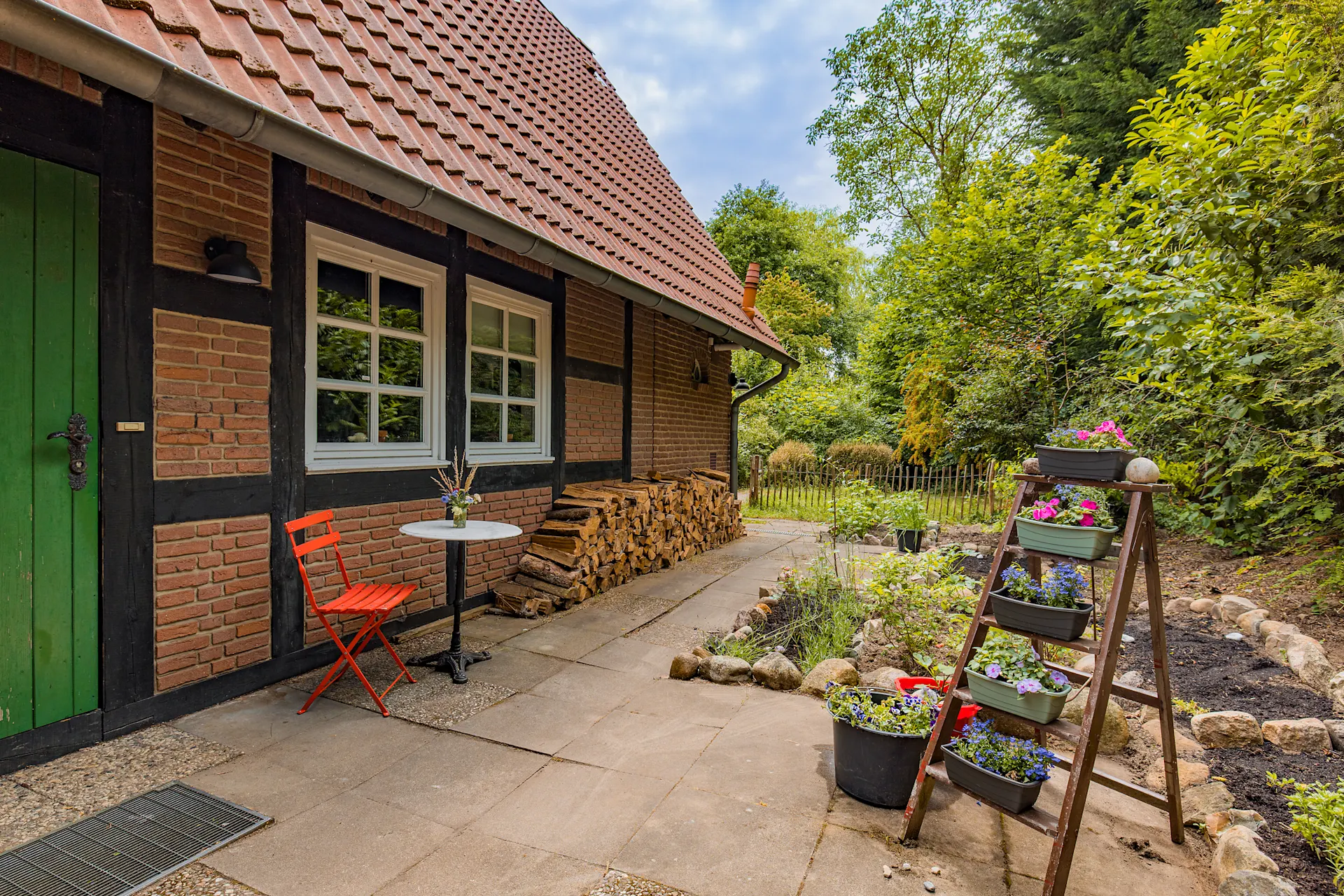 Garten mit rotem Stuhl, kleinem Tisch, Holzstapel und Blumen auf einer Leiter neben einem Backsteinhaus.