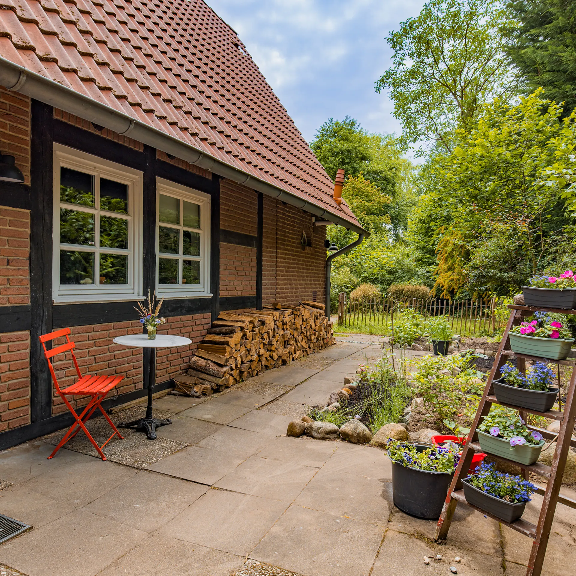 Garten mit rotem Stuhl, kleinem Tisch, Holzstapel und Blumen auf einer Leiter neben einem Backsteinhaus.