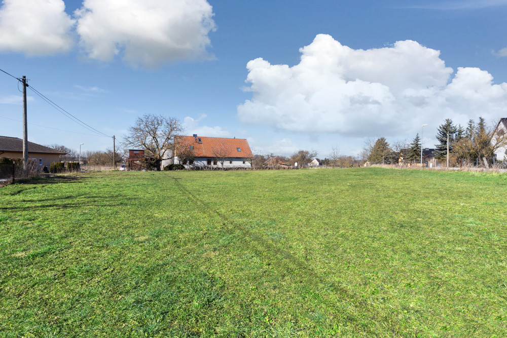 Grünes Grundstück mit Rasen, im Hintergrund stehen Wohnhäuser und Bäume unter blauem Himmel.