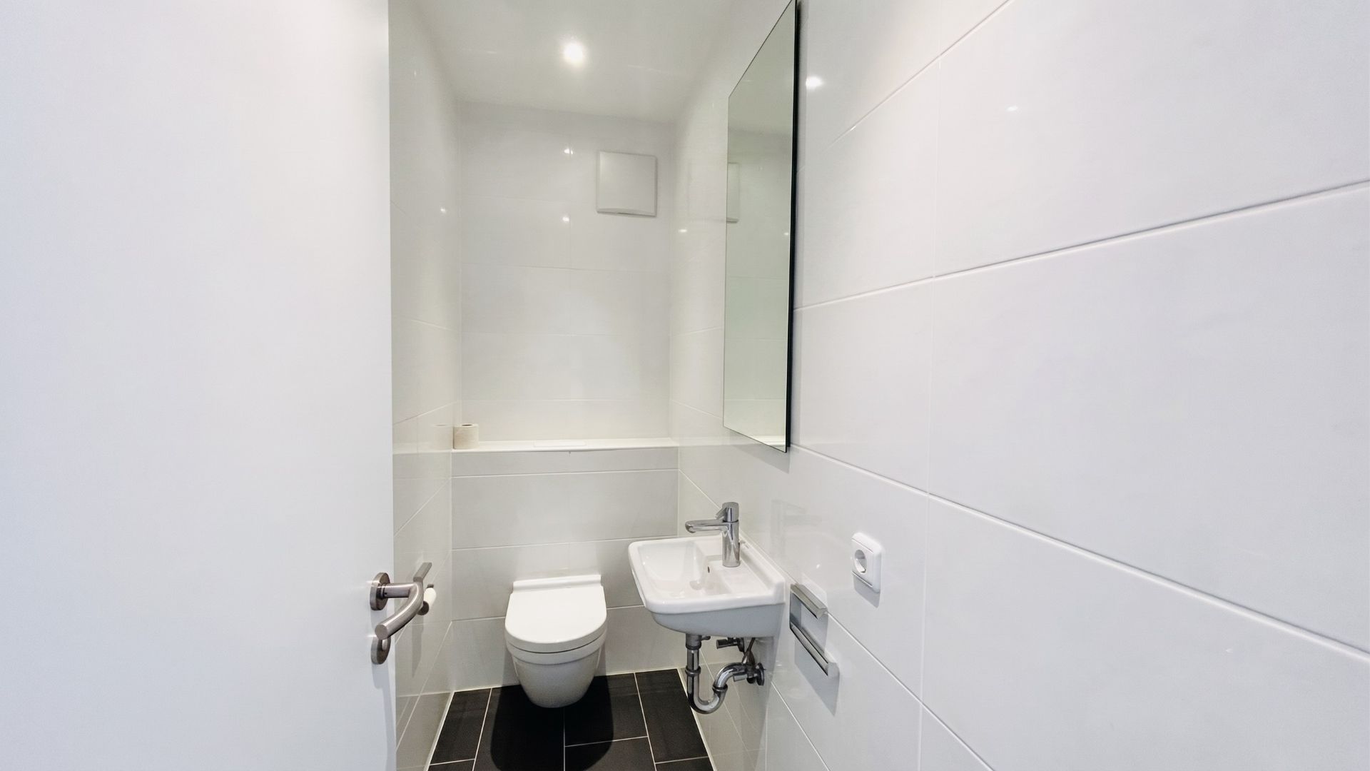 Kleines Badezimmer mit wandmontiertem WC, Waschbecken und Spiegel, weiße Fliesen, dunkler Boden.