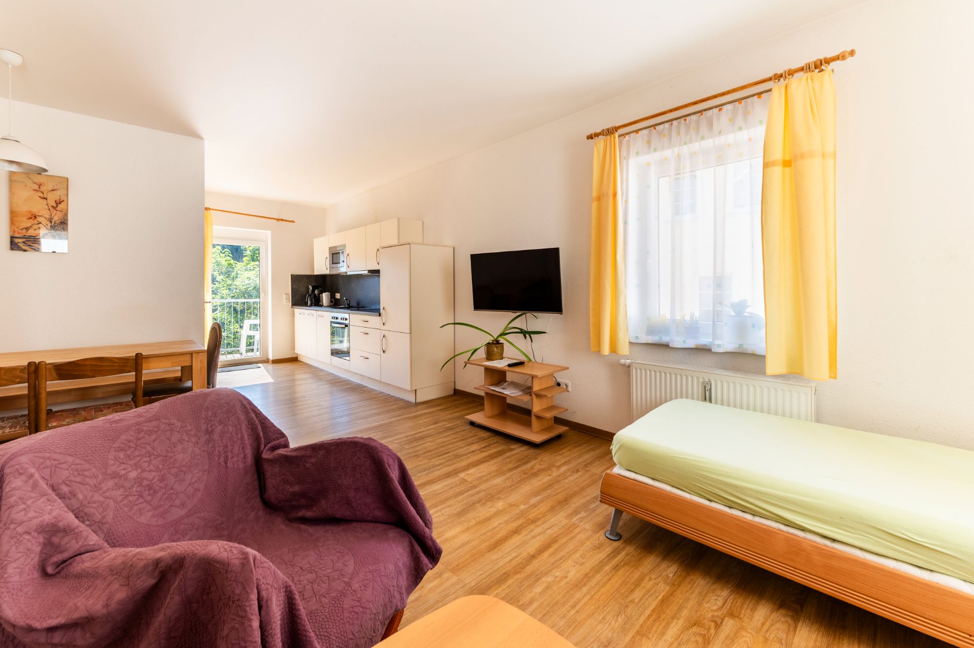 Helles Wohnzimmer mit Couch, Einzelbett, Esstisch, Fernseher und offener Küche, Fenster mit gelben Vorhängen.