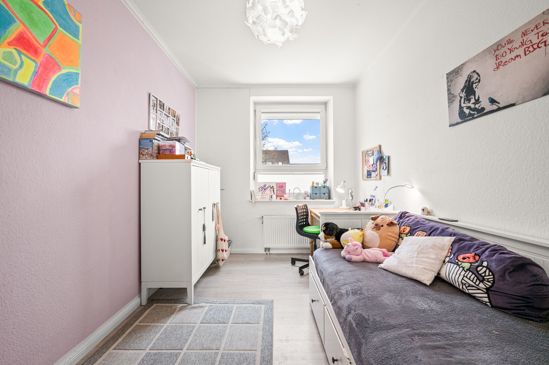 Helles Kinderzimmer mit Bett, Schreibtisch am Fenster, Schrank und Wanddekoration.