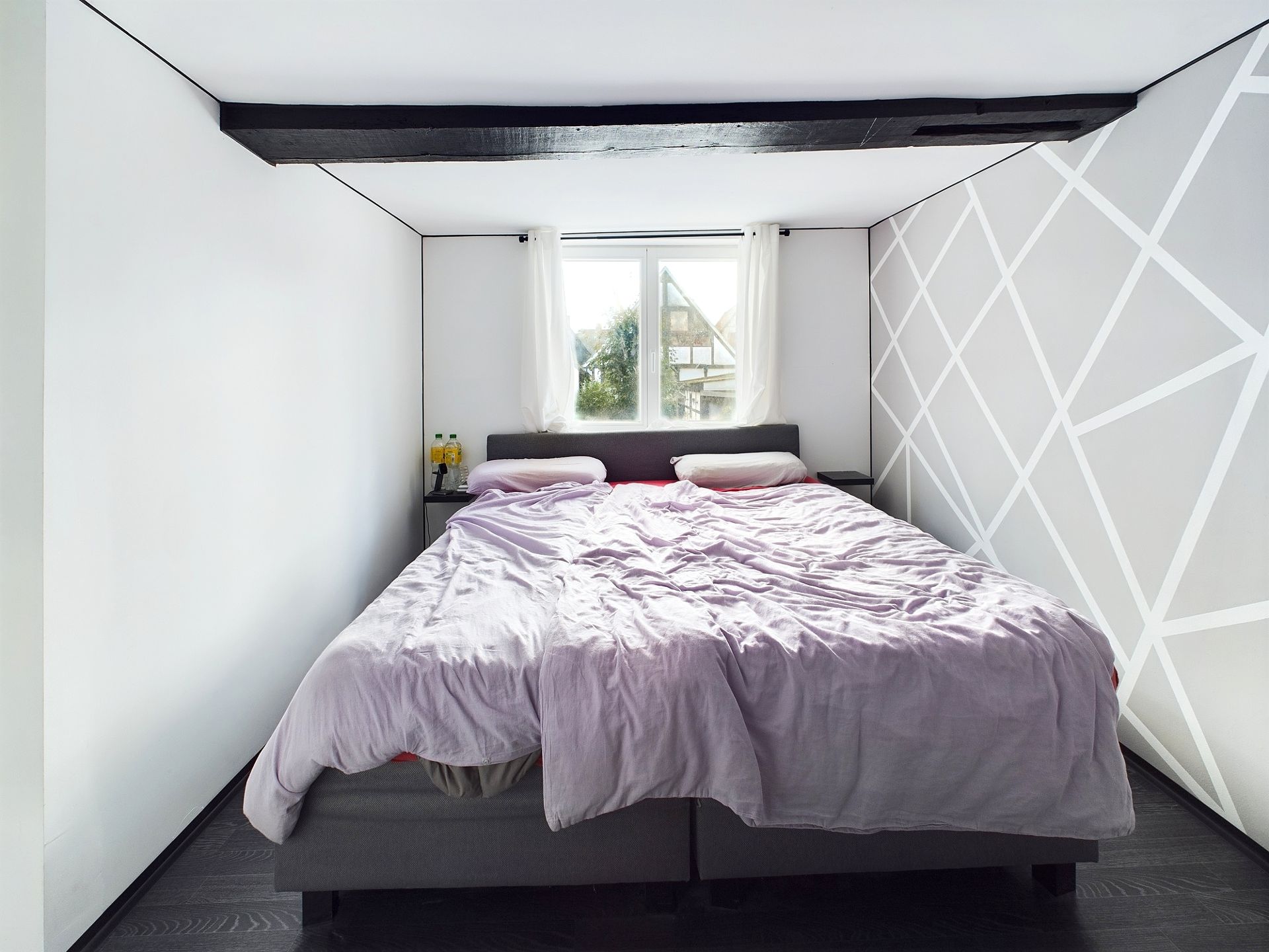 Schlafzimmer mit Doppelbett, weißer Wand mit Linienmuster und Fenster mit Vorhängen.