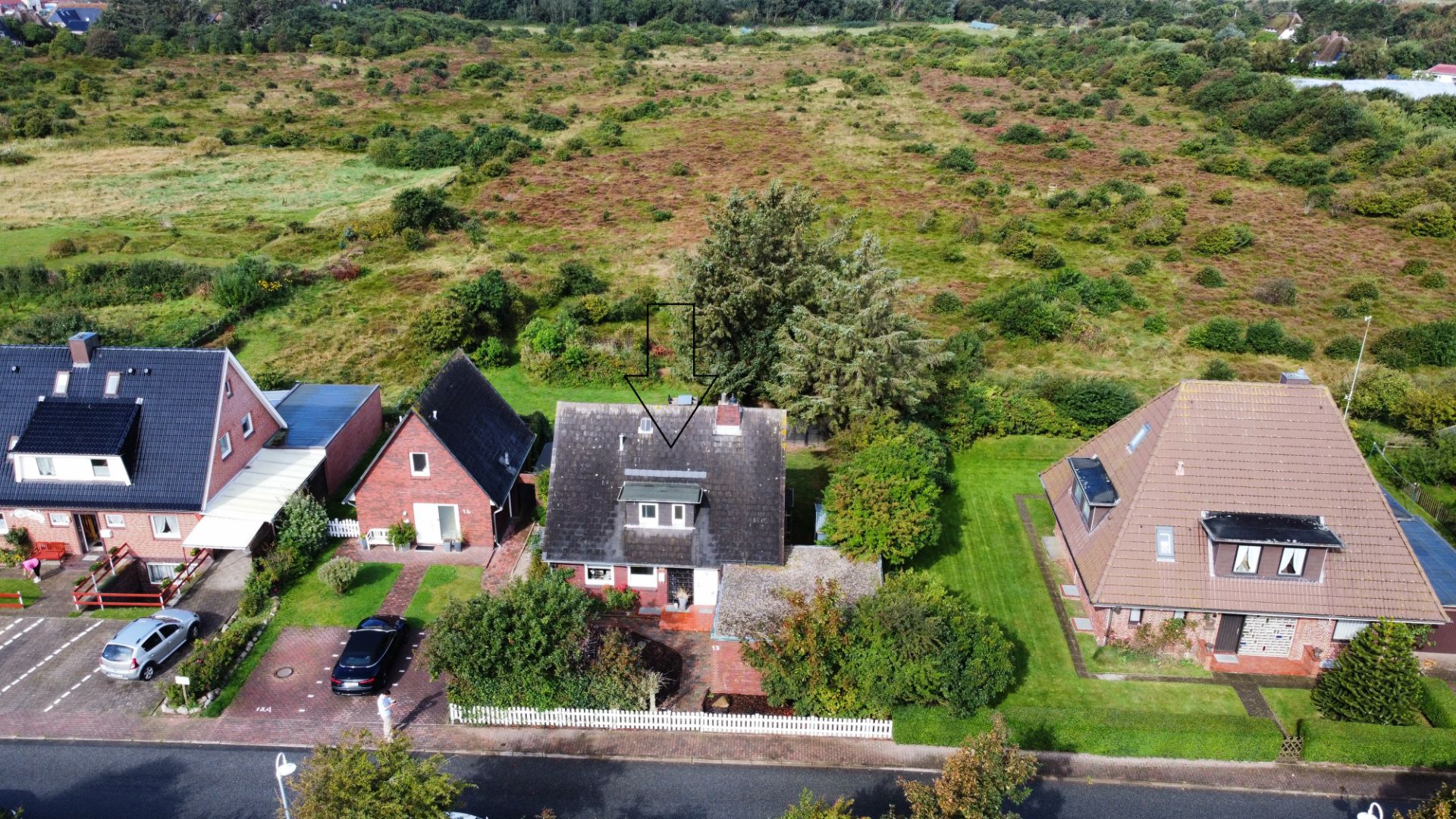 Luftaufnahme von drei Einfamilienhäusern mit Gärten an einer ruhigen Straße vor einer großen Wiese.