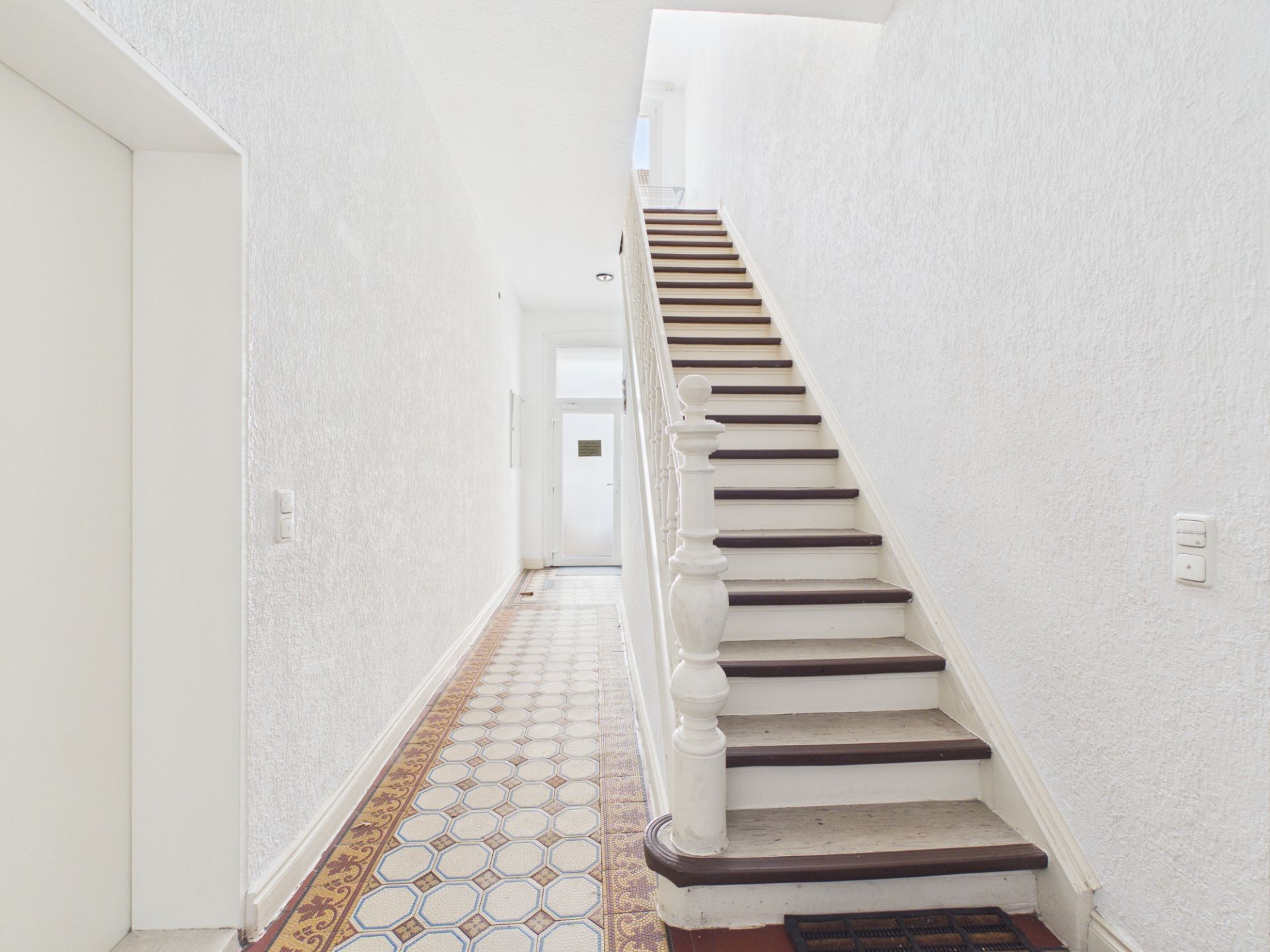 Heller Flur mit gemustertem Boden und weißer Holztreppe in einem Wohnhaus.