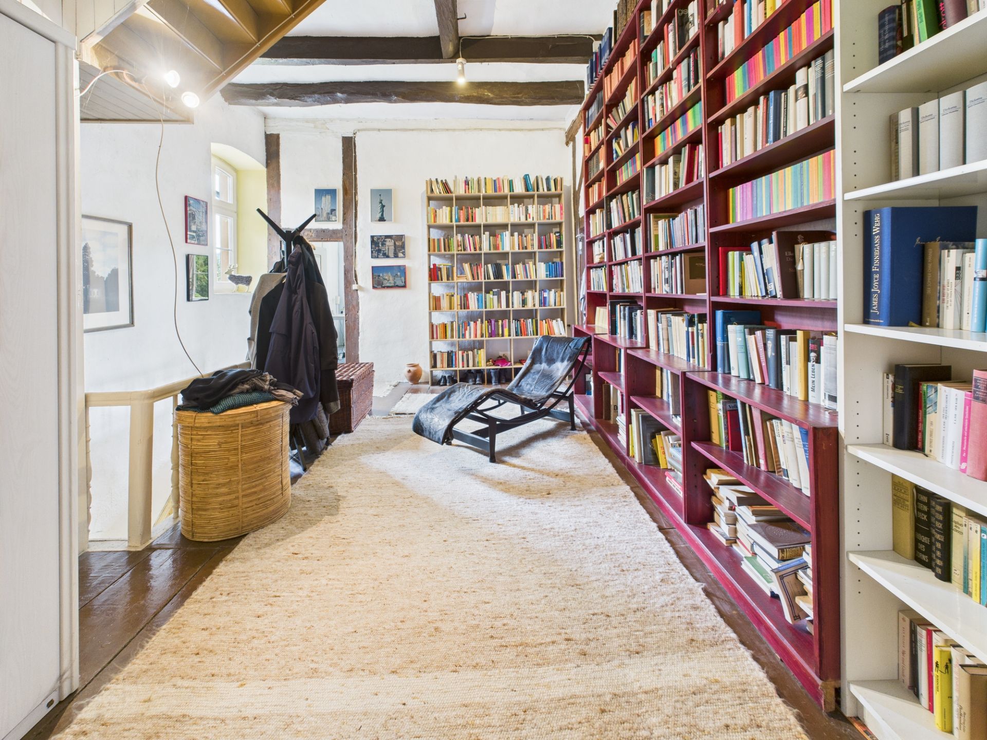 Heller Raum mit vielen Bücherregalen, einem Liegestuhl und einem Teppich auf Holzboden.