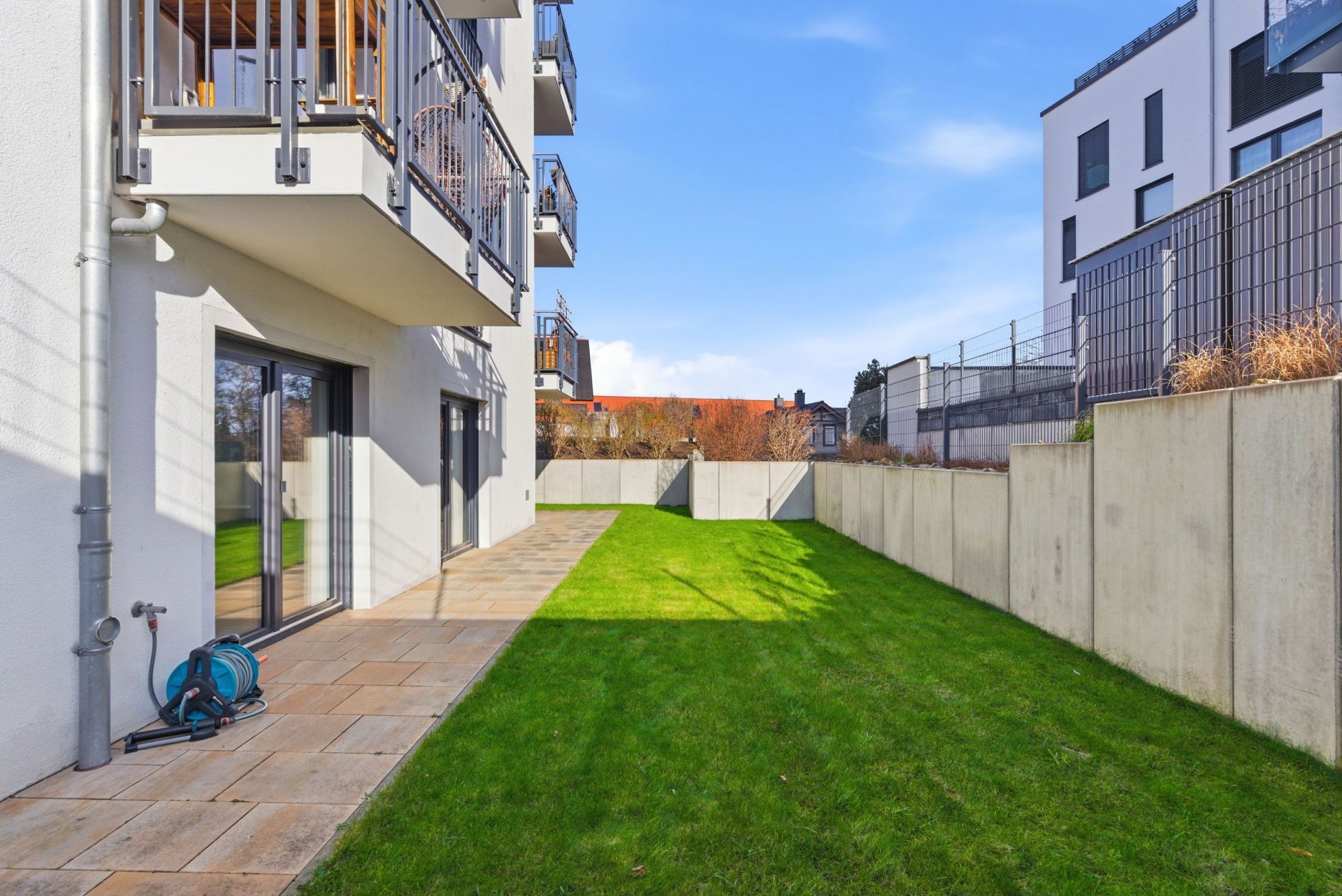 Garten mit Rasen, Terrasse und Balkonen an einem modernen Wohnhaus, umgeben von Mauern.